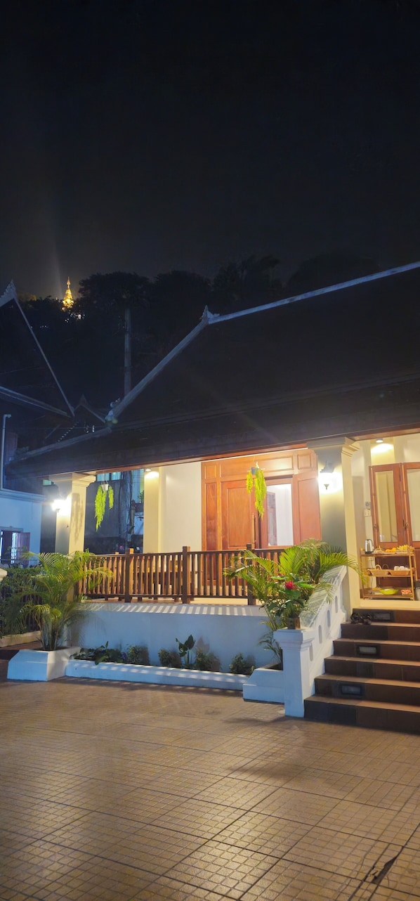 A building with a wooden balcony, steps leading to the entrance, and a view of a hill with a lighted structure in the background. - Loaviengluabgprabang guesthouse (Luang Prabang)