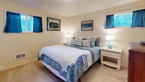 Un dormitorio con cama, mesitas de noche, cómoda y dos ventanas con cortinas.
