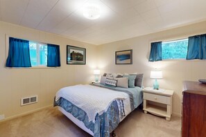 Un dormitorio con cama, mesitas de noche, cómoda y dos ventanas con cortinas.