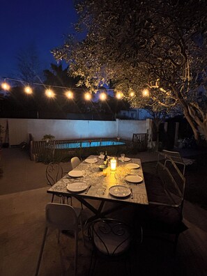 An outdoor dining area with a table set for four, surrounded by chairs, under string lights. - Very pleasant house with swimming pool, garden, air conditioning - 10 min from the beaches (Pérols)