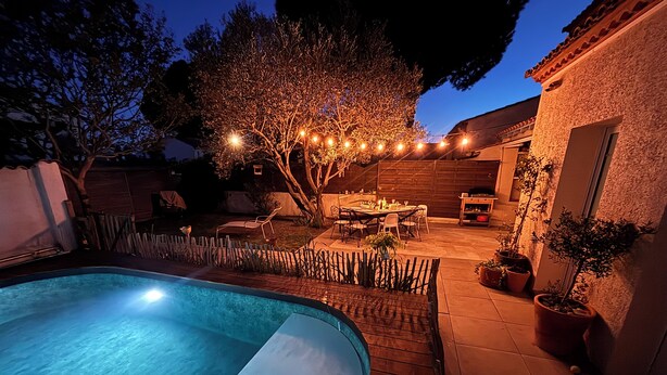 Ein Schwimmbecken mit Holzterrasse, ein Essbereich mit Stühlen und Tisch sowie ein Baum mit Lichterketten.