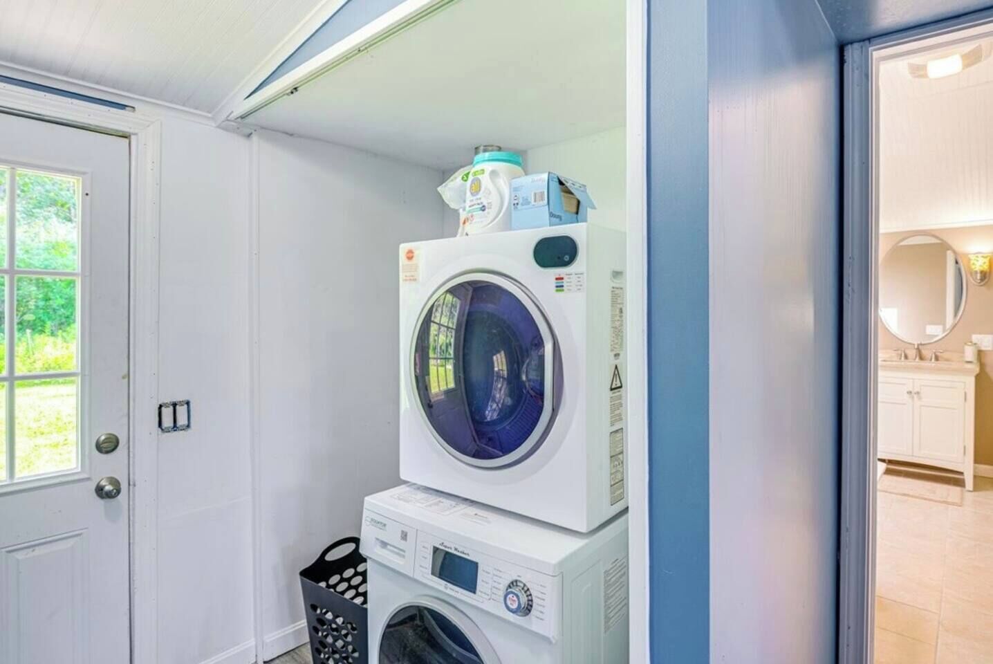A laundry area with a stacked washer and dryer, a door, and a view into a bathroom with a sink and mirror.