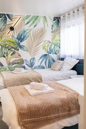 Une chambre à coucher comprenant un lit, une table de chevet, un mur orné d’un motif de feuilles tropicales et une fenêtre avec des rideaux.
