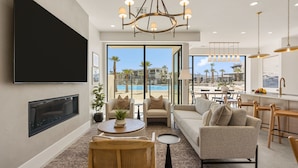 A modern living room with a flat-screen TV, a chandelier, a sofa, and a dining area with a view of a pool. - Barefoot Beach Bungalow 9 Bedroom Home (St. George)