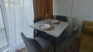 A dining area with a table set for two, chairs, a radiator, and a window with curtains. - Roma Apartment luxury flat in  Mamaia Beach by the Black Sea (Năvodari)