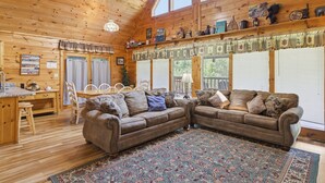 A cabin interior with wooden walls, a large window, a ceiling fan, a dining area, and a living room with a sofa and chairs. - Hibernation Station 2 Bedroom Cabin (Sevierville)