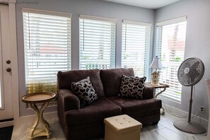 Ein Wohnzimmer mit einer Couch, zwei gemusterten Kissen, einem kleinen runden Tisch, einem Standventilator und einer Lampe.