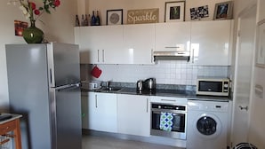 Kompakti keittiö, jossa on valkoiset kaapit, ruostumattomasta teräksestä valmistettu jääkaappi, mikroaaltouuni, leivänpaahdin, vedenkeitin ja pesukone.