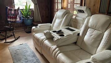 A beige leather sofa with a built-in tray table and remote controls, a rocking chair, a blue potted plant, and a window with wooden blinds.