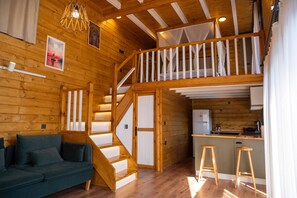 Interno di una baita in legno con divano, scala, angolo cottura e un balcone con un letto.