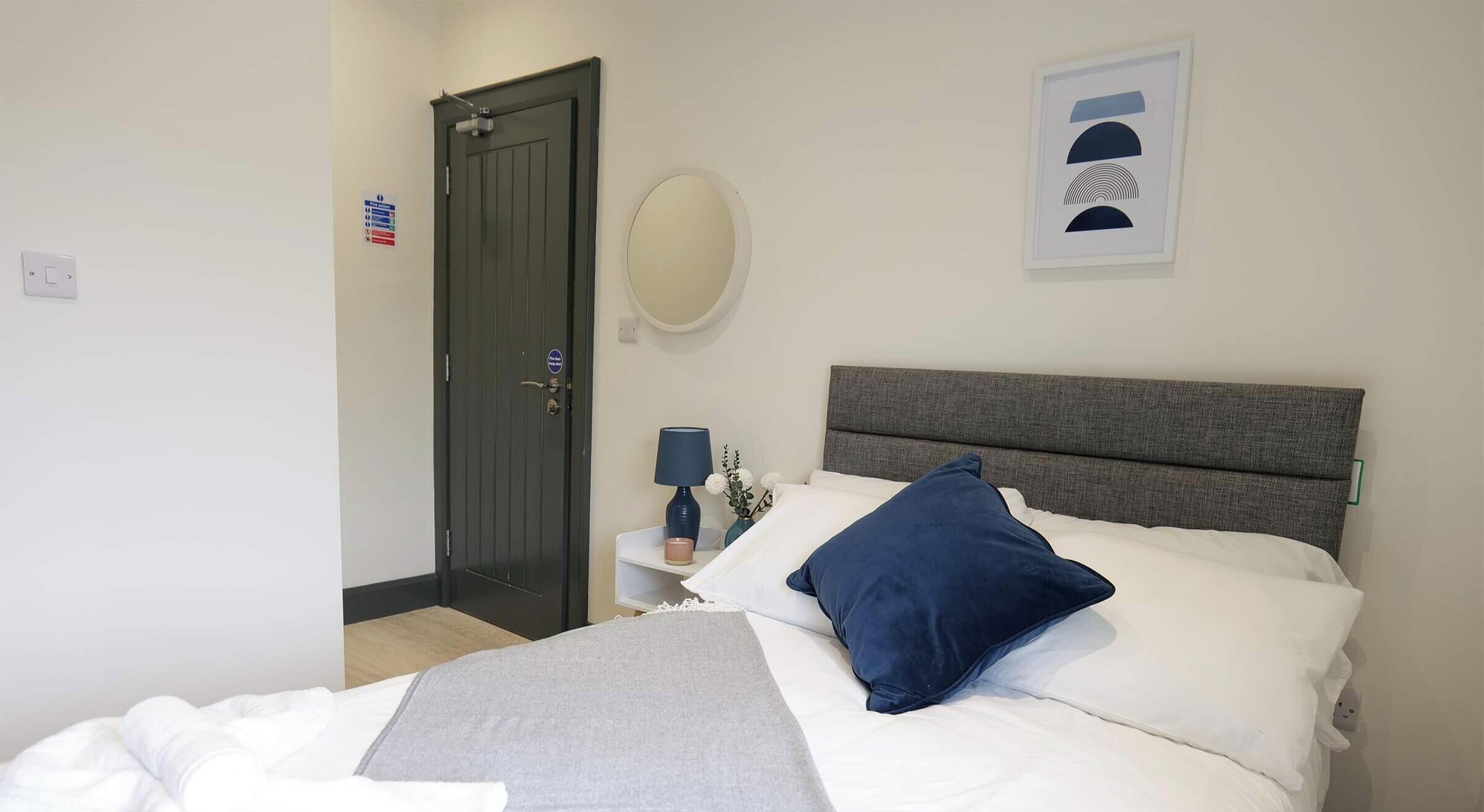 Een hotelkamer met een bed, nachtkastje met een lamp, een spiegel en een deur.