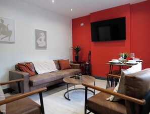 A modern living room with a sofa, coffee table, and chairs, a dining area with a table set for two, and a flat-screen TV mounted on the red wall. - Private Double Room, Shared Bathroom-Walk to City (Derry/Londonderry)