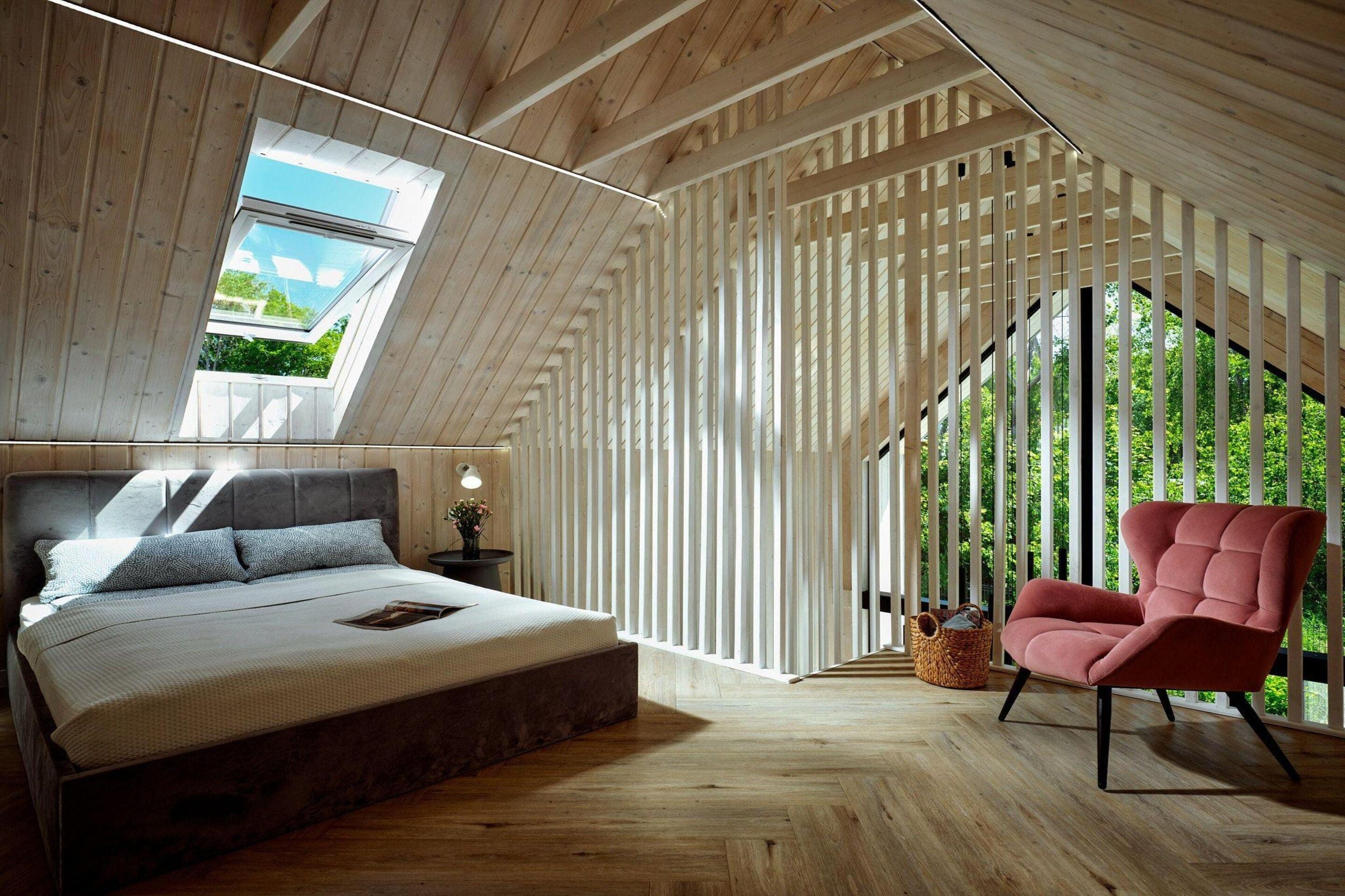 A bedroom with a sloped ceiling, a bed, a chair, and a window with a view of greenery.