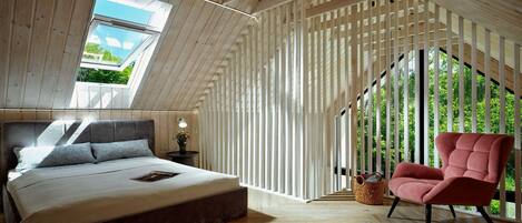 A bedroom with a sloped ceiling, a bed, a chair, and a window with a view of greenery.