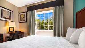 Habitación de hotel con cama, escritorio con silla, lámpara, ventana con cortinas y cuadros en la pared.