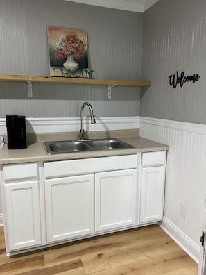 Et køkken med en dobbeltvask, hvide skabe, en træhylde og et skilt med teksten 'Welcome' på væggen.