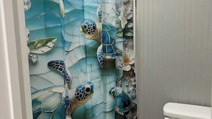 Une salle de bain avec un rideau de douche en mosaïque représentant des tortues de mer et des fleurs, des toilettes blanches et un plancher en bois.