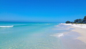 Uma praia com águas turquesa cristalinas, areal arenoso e algumas pessoas ao longe.