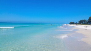 Ein Strand mit klarem türkisblauem Wasser, einem sandigen Ufer und einigen Personen in der Ferne.