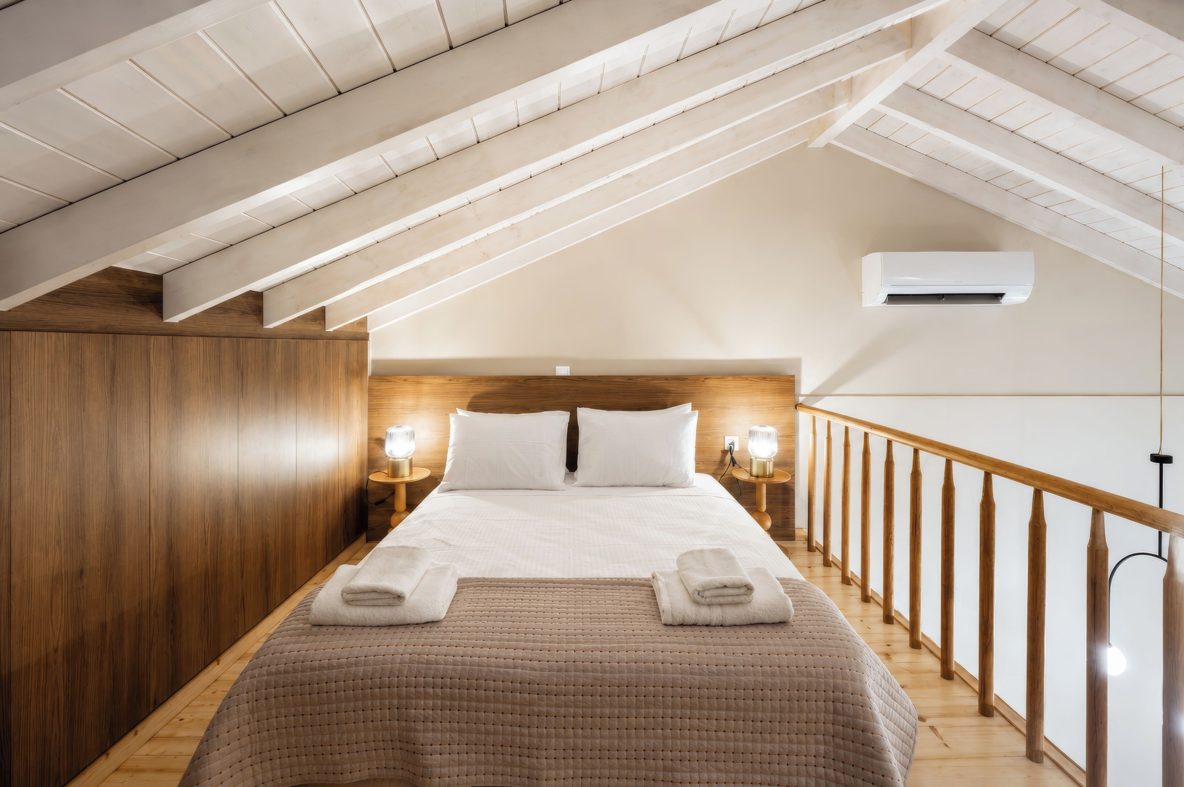 A bedroom with a sloped ceiling, wooden walls, a bed with white linens, two bedside lamps, and a wooden railing along the window.