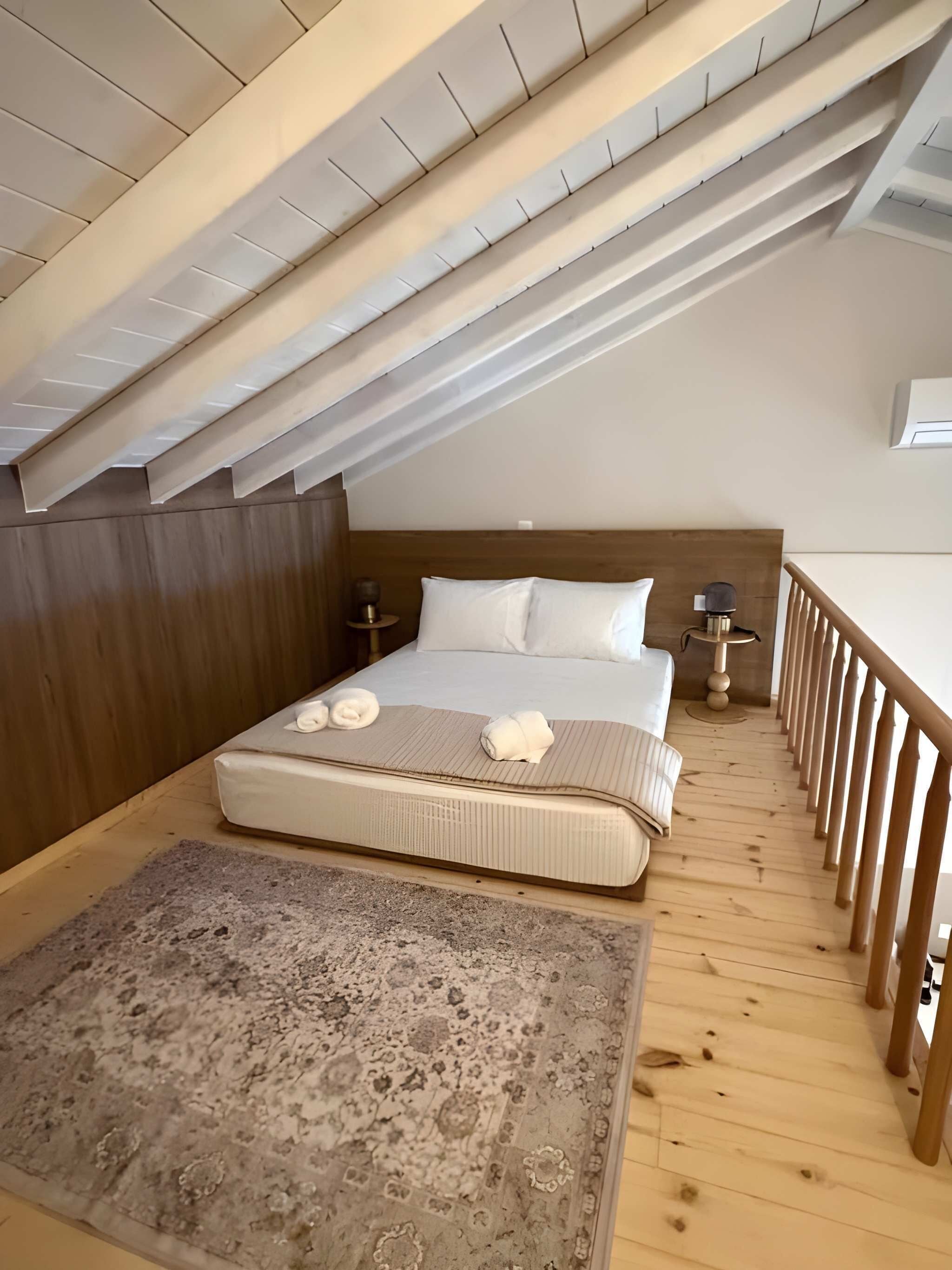 A bedroom with a wooden ceiling, a bed with white bedding, two bedside tables with lamps, and a wooden railing along the window.