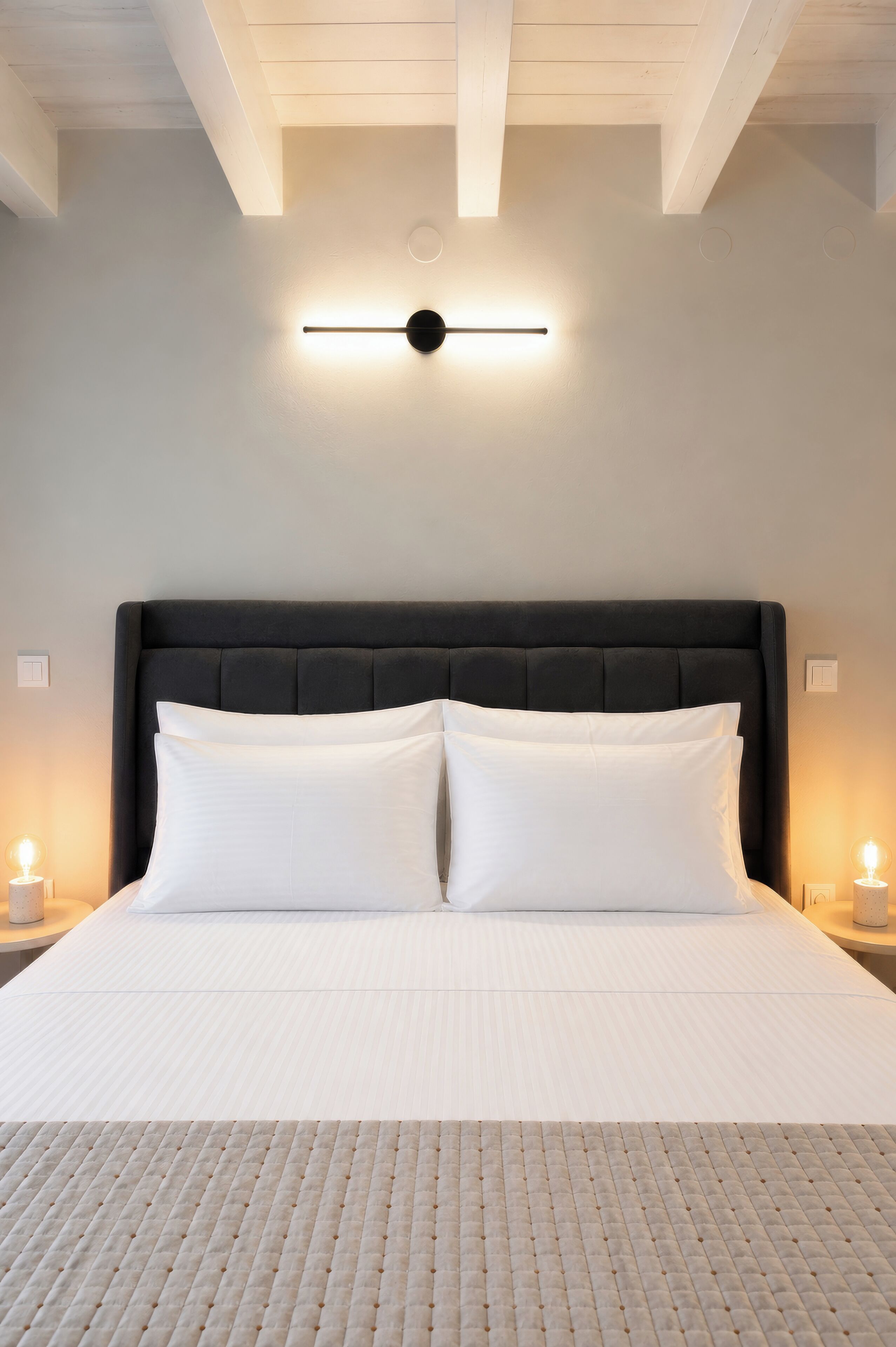 Una cama bien hecha con cabecera, dos lámparas de noche y una lámpara fijada a la pared.