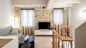 A living room with a stone wall, a sofa, a TV on a white cabinet, a wooden staircase, and a black coffee table. - Laena Apartment by All About Paxos (Gaios)