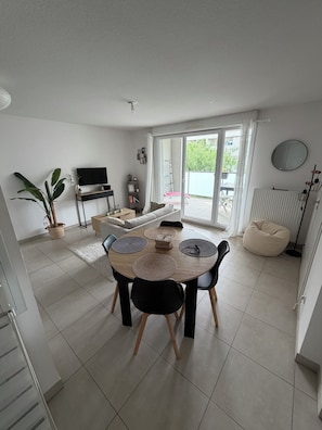 A modern living room with a dining area, a TV, a plant, and a sliding glass door leading to an outdoor area. - T2 with Balcony & Parking | 12 min from Centre (Castelnau-le-Lez)