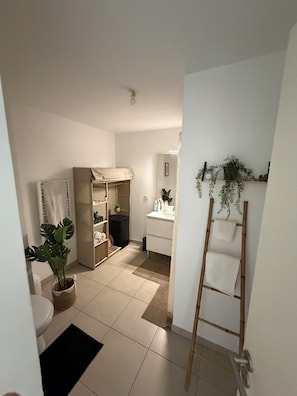 A modern bathroom with a toilet, sink, and shower area. There is a potted plant, a towel rack, and a wooden ladder leaning against the wall. - T2 with Balcony & Parking | 12 min from Centre (Castelnau-le-Lez)