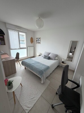 A bedroom with a bed, a desk, a chair, a small table with a plant, a radiator, a window with curtains, and a wall-mounted mirror. - T2 with Balcony & Parking | 12 min from Centre (Castelnau-le-Lez)