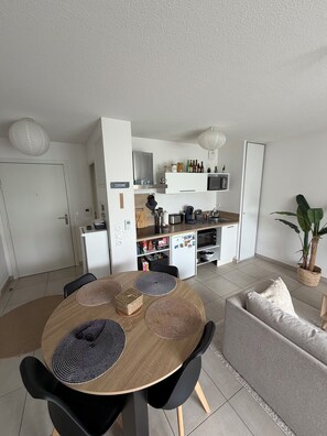A modern kitchen with white cabinets, a microwave, and a coffee machine. There is a dining table with black chairs and a potted plant. - T2 with Balcony & Parking | 12 min from Centre (Castelnau-le-Lez)