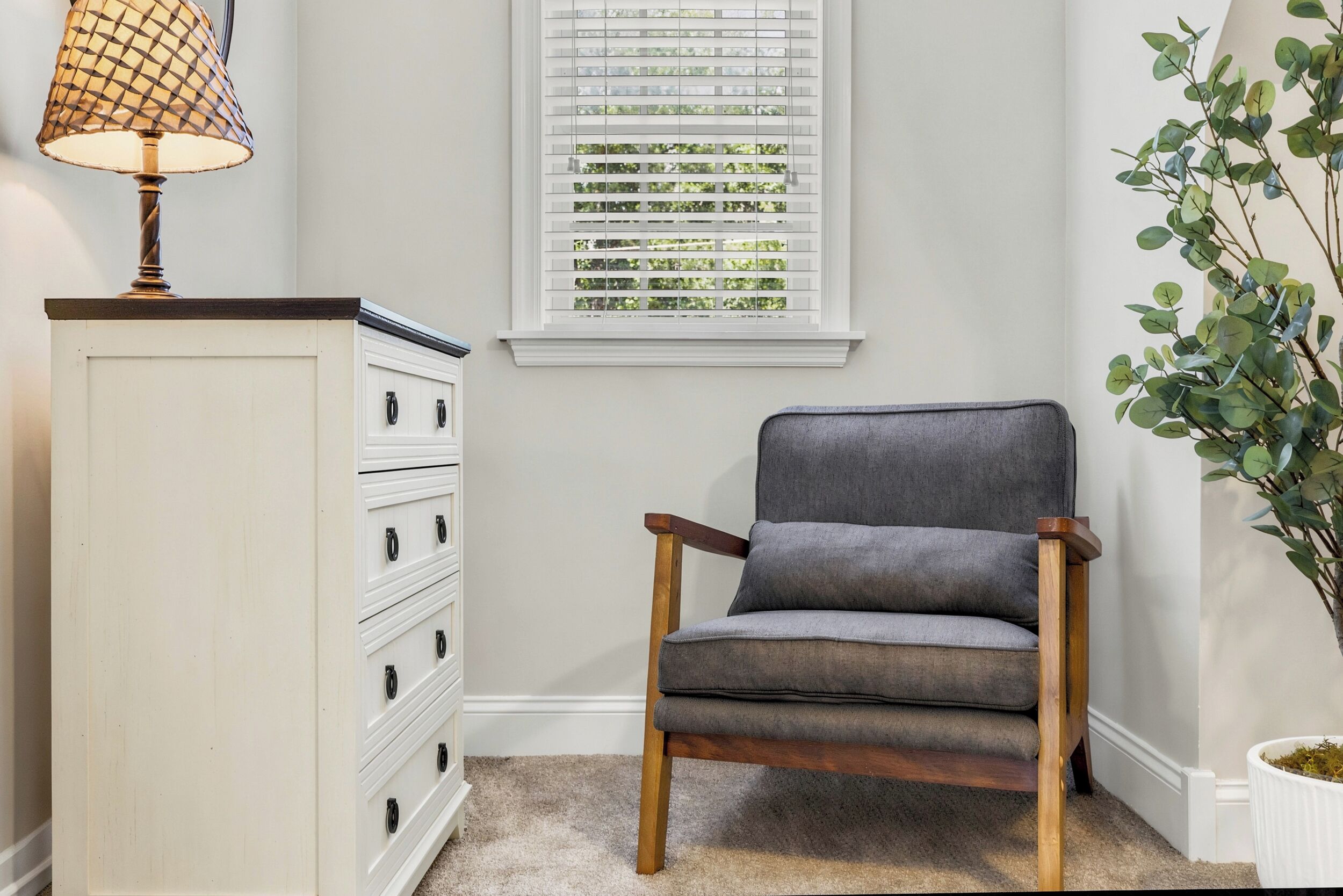 A chair, a lamp, a window with blinds, a plant, and a white dresser.
