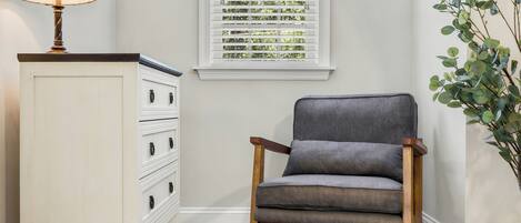 A chair, a lamp, a window with blinds, a plant, and a white dresser.