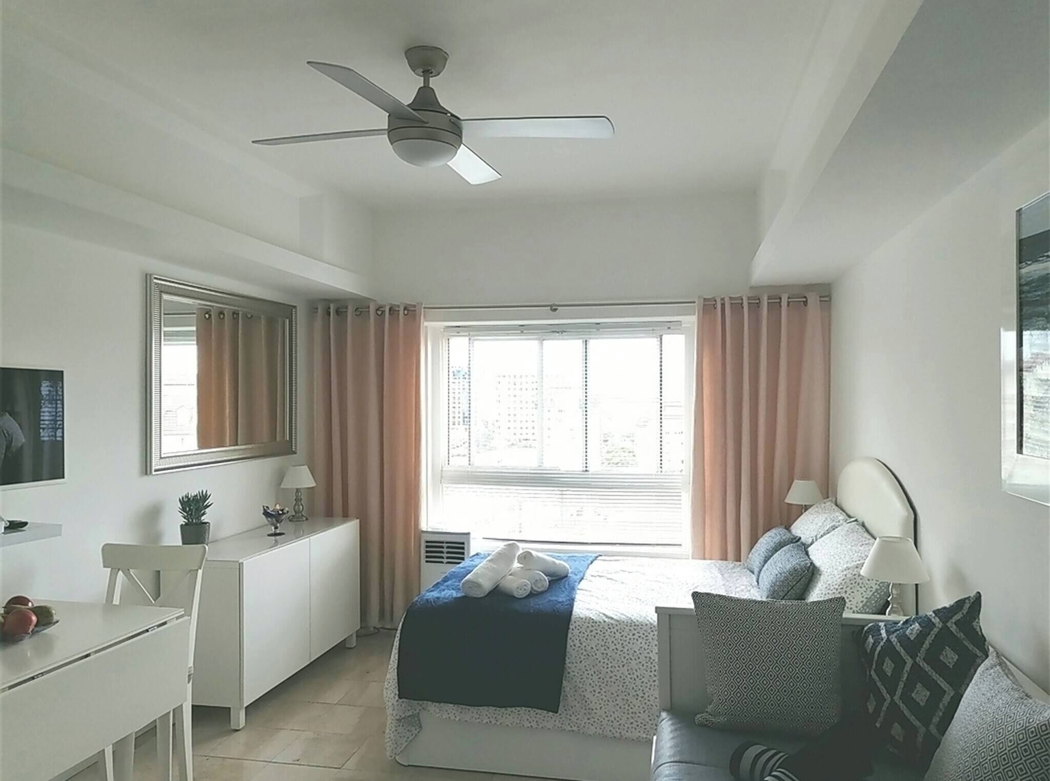 A bedroom with a bed, a desk, a chair, a ceiling fan, a window with curtains, and a mirror.