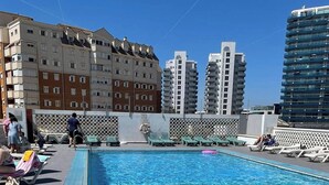 Una piscina en la azotea con sillas de descanso, zona de estar junto a la piscina y edificios altos al fondo.