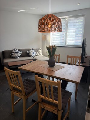 A dining area with a wooden table, chairs, a hanging light fixture, a sofa with cushions, a floor lamp, and a television. - RS Home CB 1 - Stylish apartment for vacation & business (Cottbus)