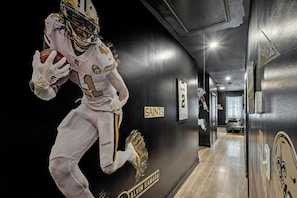 Un pasillo con un jugador de fútbol americano con uniforme blanco, un cartel que dice 'SAINTS' y una puerta con la leyenda 'RESERVES'.