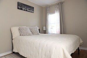 A bedroom with a bed, pillows, a nightstand, a lamp, and a window with curtains. - 3-bedroom house in Tampa near USF & Bush Gardens - Fenced Yard (Tampa)