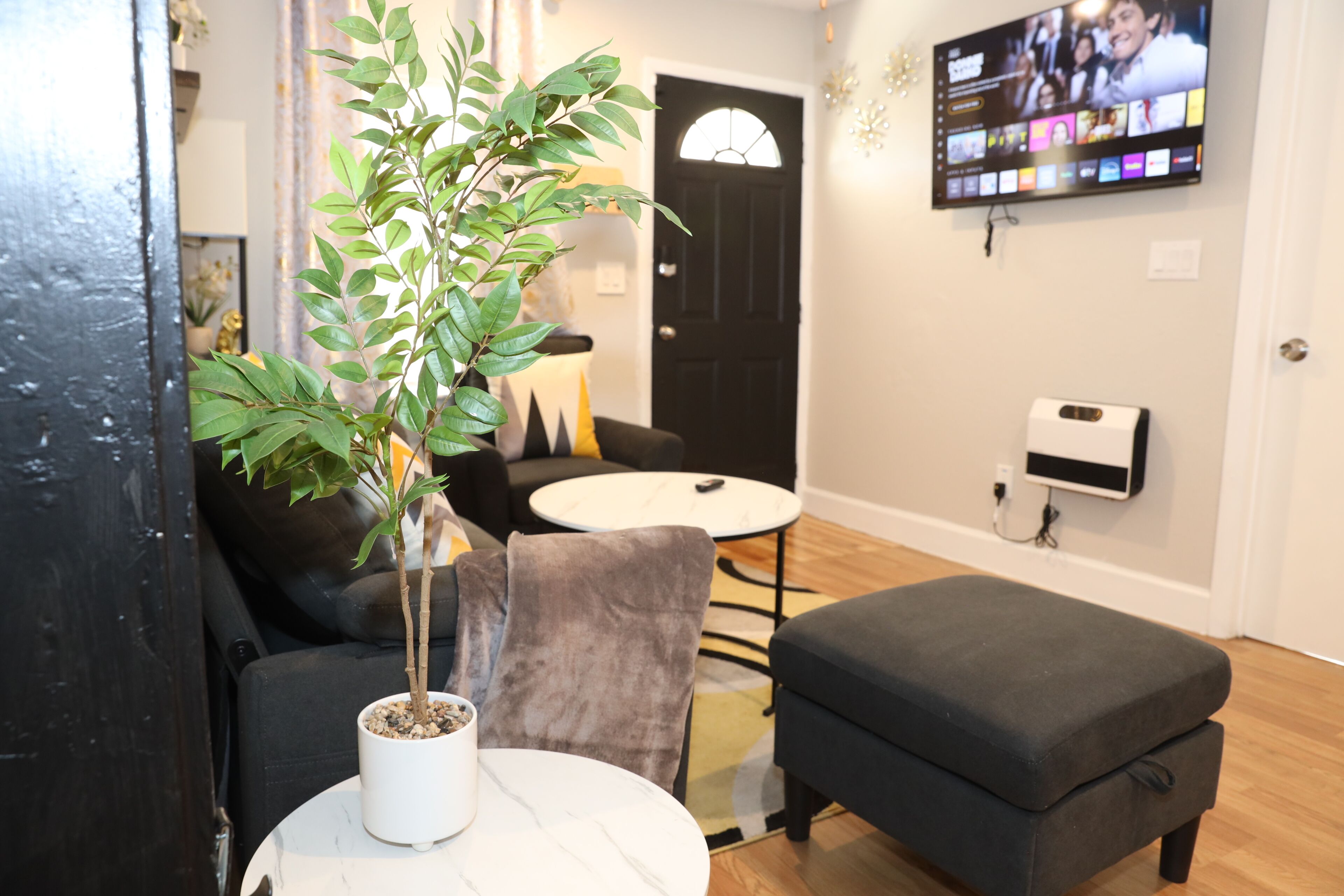 Ein Wohnzimmer mit einer Couch, einem kleinen runden Tisch, einer Topfpflanze und einem an der Wand befestigten Fernseher.