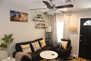 Un salon avec un ventilateur de plafond, un tableau représentant un coucher de soleil, un canapé agrémenté de coussins à motifs, une petite table et une plante en pot.