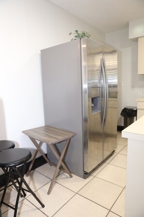 A compact kitchen with a stainless steel refrigerator, a small wooden table, and two black chairs. - 3-bedroom house in Tampa near USF & Bush Gardens - Fenced Yard (Tampa)