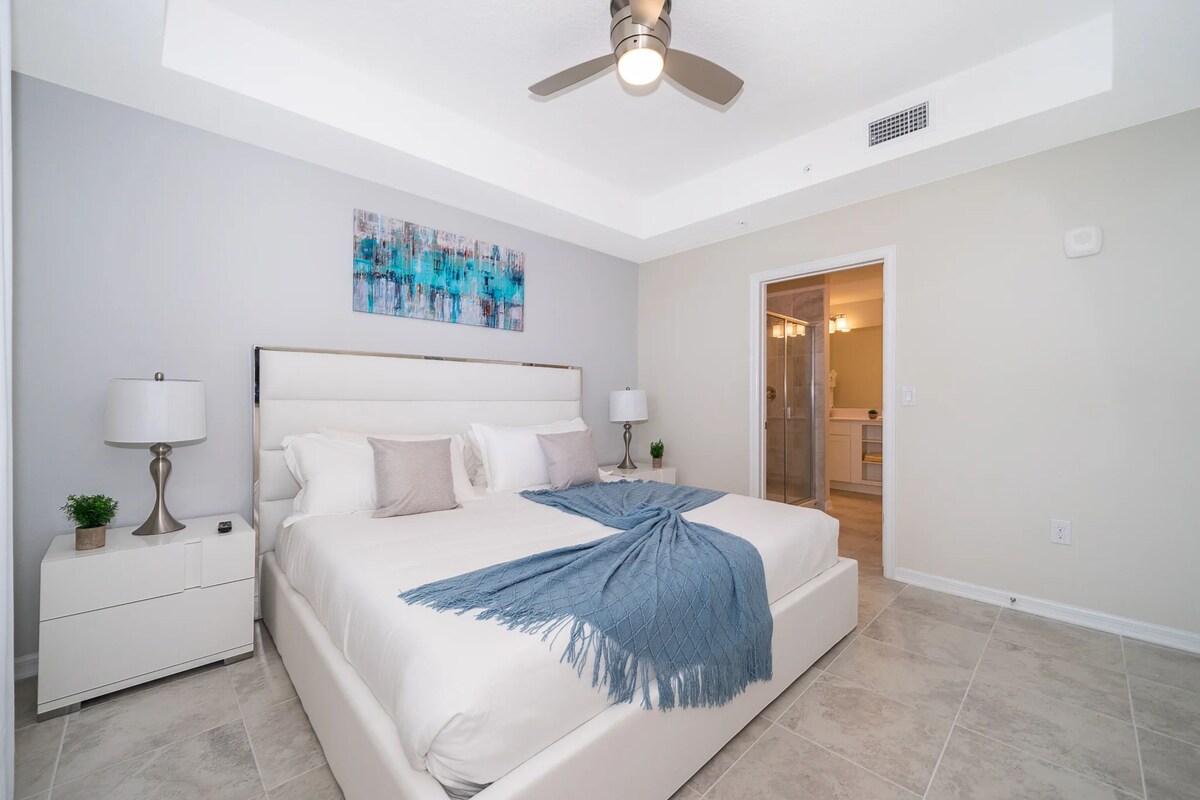 A bedroom with a bed, nightstands, a ceiling fan, a painting, and a bathroom door.