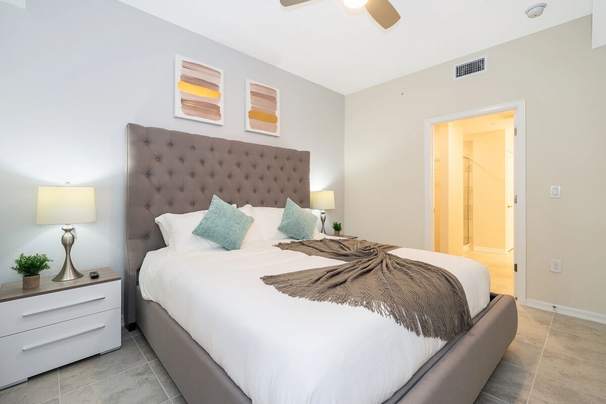 A neatly made bed with a tufted headboard, two bedside lamps, and a door leading to a bathroom.