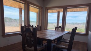A dining area with a large wooden table and six chairs, large windows with wooden frames, and a view of a natural landscape. - Mountain Top Sunsets on 12 secluded acres!  Brand new house finished in 2026 (Mena)