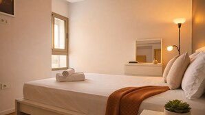 Una cama bien hecha con edredón blanco y una toalla enrollada, una mesita de noche con planta en maceta, una lámpara en el buró y una ventana con persianas.