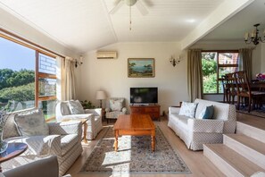 Un salon spacieux doté d’une grande télévision, de sièges confortables, d’un coin repas et offrant une vue sur la verdure environnante.