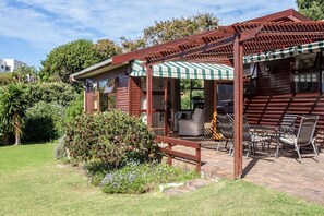 A wooden cabin with a covered patio, a small garden, and a view of the surrounding greenery. - Views, Beaches, Barbeques and more at Tom's Cabin: 3 Bedrooms, Sleeps 6 (Cape Town)