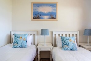 Two beds with matching headboards and nightstands, each with a lamp and a pillow with a sea-themed design.