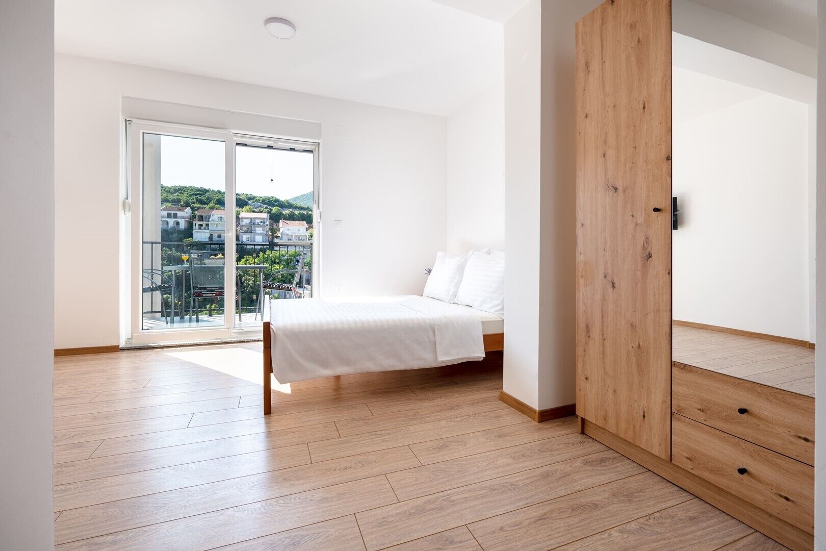 Quarto com cama, guarda-roupa de madeira e varanda com vista para casas.