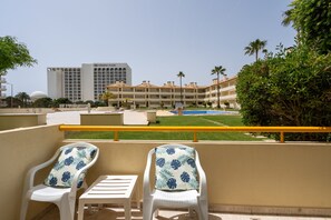 Ein Balkon mit zwei weißen Stühlen und einem kleinen Tisch, mit Blick auf einen Swimmingpool und ein Hotelgebäude.